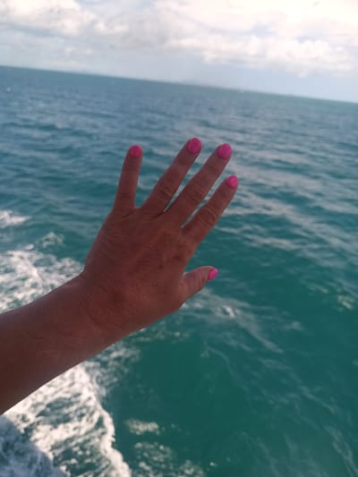 A woman's hand extends over a body of water. The water is turquoise with white foam from the boat's movement. The woman's fingernails are painted a bright pink. The location is likely Thong Sala, Thailand, based on the turquoise water and tropical appearance. The sky is visible in the background.