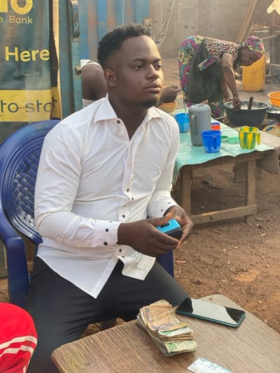 A man sits on a blue chair, wearing a white button-down shirt and black pants. He holds a blue mobile phone. A stack of paper money and another smartphone are on the wooden table. In the background, a woman wearing a colorful dress and headscarf is sitting on a stool, possibly working with water containers and buckets. A yellow advertisement is on a structure near the man.