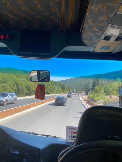 The image, taken from inside a bus, shows a view through the windshield. A digital clock displays "6 25". A black screen is mounted above the windshield. The bus interior features a patterned fabric ceiling and overhead compartments. A rearview mirror hangs from the ceiling, reflecting the road ahead and a portion of the bus's interior. A red flag with a yellow crest, likely the flag of Montenegro, hangs from the mirror.

Outside, the bus is on a paved road surrounded by lush green hills and trees. The sky is clear and blue. Several cars are visible on the road, including a silver SUV with "SORENTO" written on its side and a black sedan. Construction work is evident along the roadside, with orange fencing and piles of dirt. A sign with Cyrillic text, possibly indicating a route or destination, is partially visible on the back of a seat. The location is identified as Prijeradi, Montenegro.