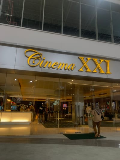 This image captures the entrance of a "Cinema XXI" movie theater, likely located within a modern shopping complex in Samarinda, Indonesia. The dominant feature is the large, three-dimensional sign with the name "Cinema XXI" displayed in gold-colored lettering on a grey facade. The word "Cinema" is in a flowing script font, while "XXI" is in a bold serif style.

In front of the cinema entrance are clear glass doors, reflecting the interior lighting and a partial view of what lies beyond. A person, wearing a light-colored top, brown trousers, and a patterned headscarf (hijab) with a dark shoulder bag, stands facing these doors, positioned on a dark green doormat. Their posture suggests they are either entering or waiting to enter the cinema.

Through the glass, reflections show parts of the cinema lobby, including glowing screens that appear to display movie information or advertisements. Other faint reflections of people and interior structures are also visible. On the glass doors, the text "Cinema XXI" is visible in reverse due to reflection, alongside a "no entry" or "no specific item" symbol. To the left of the cinema entrance, a large windowed section of the building reveals internal support structures and what appears to be some scaffolding. The scene is brightly lit by artificial indoor lighting, giving no clear indication of the time of day beyond being an active commercial environment.