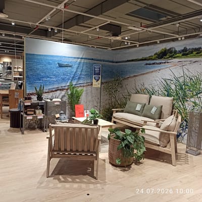 This image depicts an indoor furniture store, likely part of a chain, showcasing outdoor or garden furniture. The backdrop is a large mural of a beach scene with a small boat in the water and green foliage. In the foreground, a set of light-colored wooden outdoor furniture, including a sofa and an armchair, is arranged on a light wood-effect floor. Decorative elements such as potted plants in woven baskets, small decorative boxes, and a lantern-like fixture are also visible. A price tag reading "450€" is displayed on a small table. The lighting suggests an artificial environment typical of retail spaces. The timestamp "24.07.2026 10:00" is superimposed on the lower right of the image. There is no indication of the specific location within the city of Modugno, Italy, beyond it being an indoor retail setting.