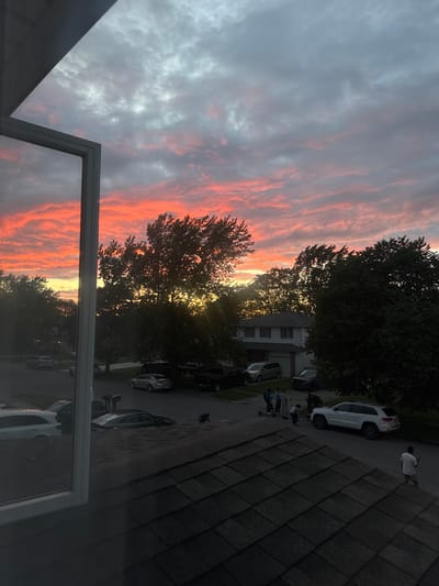 A sunset view from an elevated perspective shows a suburban street in the Town of Cheektowaga, United States of America. The sky is filled with dramatic clouds displaying hues of orange, pink, and gray. In the foreground, a portion of a window frame and a dark shingled roof are visible. The street below is lined with parked cars, including SUVs and sedans. A group of young people are gathered on the street, some with scooters. Trees with dense foliage flank the street and the houses. The scene suggests a warm, late afternoon or early evening in a residential area.