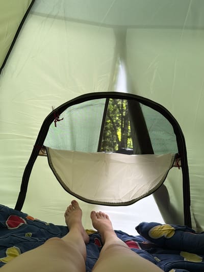 The image shows the interior of a light green tent, with the view looking outwards through an open mesh-covered entrance. The entrance is framed by black piping and is partially covered by a white fabric panel that is zipped down. Outside the tent, through the mesh, trees and foliage are visible, suggesting a natural environment, possibly a forest. In the foreground, the lower legs and feet of a person are visible, lying on a dark blue blanket with a pattern of colorful shapes. The person appears to be relaxed, with their feet resting on the blanket. The scene suggests a person camping in a wooded area. There is no visible information to suggest the location is in the city of Rozhkao, Russia.