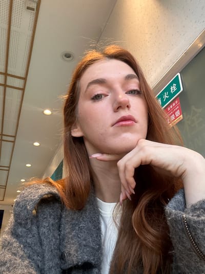 A woman with long auburn hair and fair skin poses indoors. She's wearing a grey textured sweater over a white shirt. Her hand rests on her chin. The background includes a ceiling with recessed lighting and a green sign with red text. The image is rotated, giving a skewed perspective. The focus is on her face and the pose she is striking.