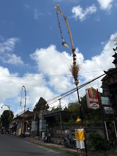 The image shows a street scene in Tampaksiring, Indonesia, with traditional Balinese decorations hanging from poles. The decorations are tall, ornate, and made of natural materials, including dried grasses and woven elements. One prominent decoration is a long, tiered structure adorned with small woven pieces and a larger bunch of dried grass at the bottom. In the background, there is a stone archway, typical of Balinese temple entrances, with carved details. Several shops line the street, with signs indicating they are "Toko" (shop). One sign reads "SINGARAJA" above "TOKO ATMAJAYA" and another is partially visible with "PHOTO COPY". The street is paved, and there are several motorcycles parked along the side. The sky is blue with scattered white clouds. The overall atmosphere suggests a cultural and possibly festive setting in a tropical environment.