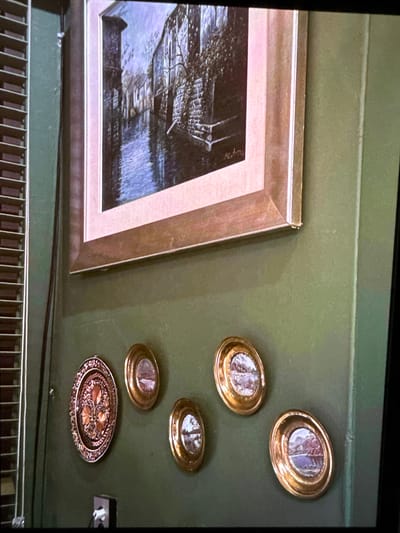 This is a photograph of a section of a wall in what appears to be an indoor setting. The wall is painted a muted green color. A large framed painting hangs on the upper portion of the wall, depicting a canal scene with stone buildings and reflections in the water. The painting has a wide, light-colored mat and a wooden frame with a subtle sheen. To the left of the painting, a portion of a window with horizontal blinds is visible. Below the large painting, a collection of smaller decorative oval objects are mounted on the wall. The largest of these is an oval piece with intricate metallic detailing, resembling a decorative plate or medallion. To its right, four smaller oval objects are arranged, each containing a miniature landscape painting within a golden frame. One of these smaller objects is slightly lower and to the right of the others. In the lower left corner of the image, a dark electrical outlet with a white plug is visible on the wall. The image does not provide any contextual information about the city of Mokpo-si, South Korea, or any people.