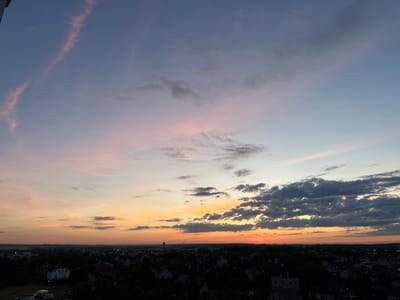 This image captures a panoramic view of a suburban landscape at dusk. The sky dominates the upper portion of the frame, transitioning from a pale blue at the top to hues of pink, orange, and yellow closer to the horizon, indicating the setting of the sun. Wispy clouds are scattered across the sky, some tinged with pink by the fading sunlight, while others are darker, more defined masses.
Below the sky, a dense residential area is visible, characterized by a multitude of houses with red, white, and gray roofs, nestled amongst numerous trees. The architecture suggests a European setting. In the distance, a tall, slender structure resembling a water tower or industrial chimney stands out against the twilight. The overall impression is one of a peaceful, quiet evening in a rural or suburban area. The dark silhouettes of trees and buildings against the brightly colored sky evoke a sense of calm and transition. The time of day is clearly sunset or dusk. There is no visible text in the image. The location context is Шчомысліцкі сельскі Савет, Belarus.