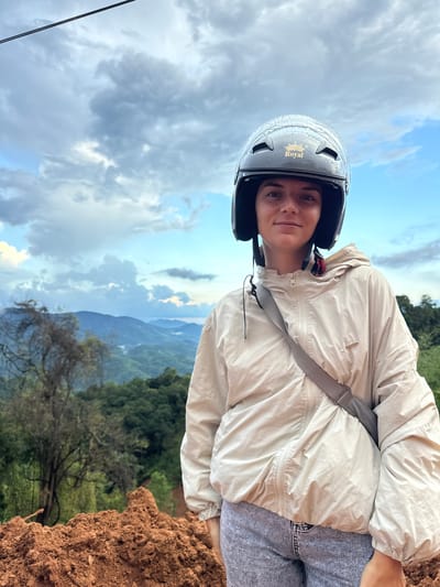 A young woman, wearing a black motorcycle helmet with a "Royal" logo and a light beige jacket, stands in front of a scenic mountain landscape. She is looking directly at the camera, with a slight smile. The background consists of lush green hills and mountains, under a cloudy sky with patches of blue. In the foreground, there is a mound of reddish-brown soil. A thin black wire is visible at the top of the frame. The location is identified as Nao Sẻ, Vietnam.