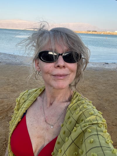 A woman with gray hair and sunglasses poses for a selfie on a beach in the Tamar Regional Council, Israel. She is wearing a red bikini top and a loose-fitting, yellow, textured cardigan with a black checkered pattern. A delicate silver necklace with a small diamond pendant rests on her collarbone.
The background shows a sandy beach leading to the calm, blue water of the sea. In the distance, hazy mountains are visible under a clear, bright sky, suggesting a sunny day, likely in the late afternoon or early evening given the soft light. The overall mood is relaxed and serene, capturing a moment of enjoyment at the seaside. There is no visible text in the image.