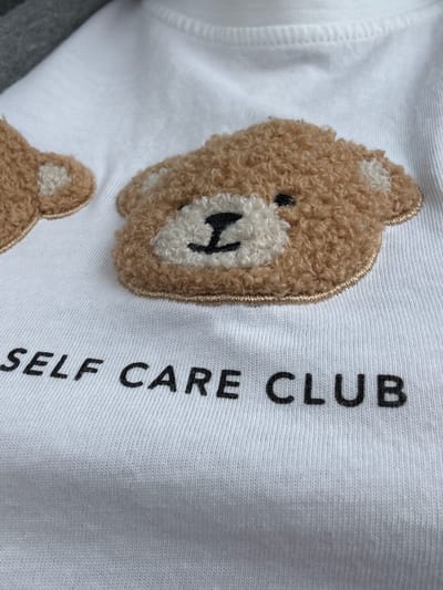 The image shows a close-up of a white t-shirt with a textured teddy bear appliqué and the words "SELF CARE CLUB" printed below it. The teddy bear is brown with white accents for its ears and muzzle, and has embroidered black eyes and nose. The text is in black, block letters. The t-shirt is made of a ribbed knit fabric. There is no indication of the city of Vellmar, Germany in the image or its context.