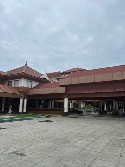 The image shows a wide, tiled plaza in front of a building with a prominent red tiled roof and brown trim. The building features a series of covered walkways supported by white columns, and the roofline has several tiered sections with decorative elements. In the background, people are visible walking and standing under the covered areas, some appearing to be staff members in traditional Indian attire. There is a patch of green grass with planters to the left of the building, and a few potted plants are visible under the covered walkways. The sky is overcast with gray clouds. The plaza appears to be an entrance area, possibly to a public building or a hotel. This appears to be the entrance to the Cochin International Airport (COK) in Kochi, India.