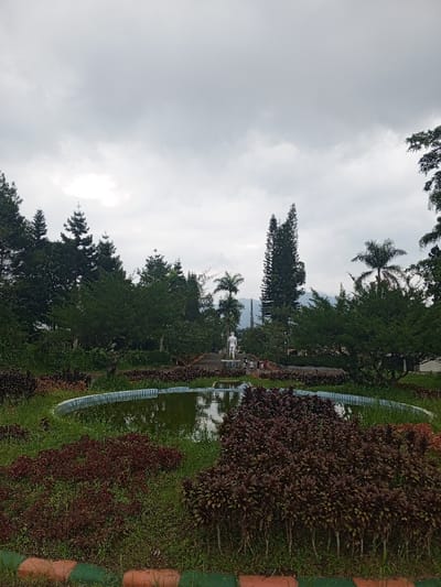 This full-shot image captures a lush, verdant park or garden scene in Baza, Myanmar, under an overcast sky. In the immediate foreground, a border with alternating orange, red, and green stripes defines the edge of the viewing frame. Beyond this, vibrant dark red and maroon foliage contrasts with green grass, leading to a large, partially circular pond or fountain. The pond contains still, greenish water, reflecting the muted sky and surrounding vegetation.
In the midground, a path leads from behind the pond towards a white statue of a standing male figure, erected on a pedestal. Several small, indistinct figures, likely people, are visible near the base of the statue, suggesting a public space. The area around the path and statue is surrounded by dense greenery, including various types of trees and shrubs. Tall, slender pine-like trees and several palm trees punctuate the skyline, interspersed with other broadleaf trees.
The background reveals a range of mountains partially obscured by the extensive tree line and the cloudy conditions. The sky is entirely covered with heavy, grey clouds, indicating an overcast day. The overall lighting is soft and diffused, contributing to a calm and somewhat somber atmosphere. No discernible text is visible in the image.