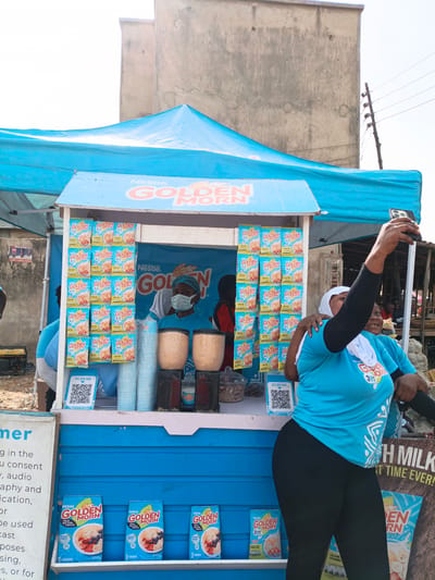 Here's a concise description of the image:
**Location:** Kawaji, Nigeria
**Scene:** An outdoor "Golden Morn" breakfast cereal stand.
**Subjects:**
* A vendor wearing a face mask is standing behind the stand.
* Two women stand on the customer side of the stand, one is holding up a smartphone, presumably taking a selfie. Both are wearing t-shirts branded with "Golden Morn" graphics.
**Objects:**
* A blue-and-white stand with "Nestle Golden Morn" branding, with packages of "Golden Morn" cereal displayed on the shelves.
* Cups and containers are displayed.
* There is additional "Golden Morn" promotional material.
* A blue canopy covers the stand.
* A QR code.
**Actions/Interactions:**
* The woman is holding up a phone, capturing a photograph.
* The vendor is standing behind the stand, presumably selling the product.
**Environment:** A busy outdoor market setting. A building is visible in the background.