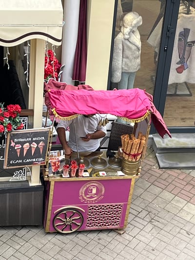 A street vendor stands behind a pink ice cream cart with a gold trim and a pink awning. The cart is adorned with decorative metalwork and a gold emblem. The vendor, wearing a white shirt, holds a mobile phone in his right hand. On the cart, there are containers of ice cream, waffle cones, and cups filled with what appears to be fruit, possibly strawberries. A chalkboard sign displays drawings of ice cream cones and Russian text. Red flowers are visible near the cart, adding a splash of color to the scene. In the background, a store window displays mannequins dressed in clothing. The scene appears to be on a cobblestone street in Kislovodsk, Russia.