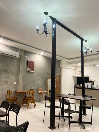 This media file depicts the interior of a modern cafe or restaurant. The scene is illuminated by artificial lighting, suggesting it could be any time of day. The setting is characterized by a blend of industrial and natural elements. A prominent feature is a large, dark, angled metal beam structure, possibly a decorative element or part of the building's architecture, which cuts across the frame.
Several tables and chairs are arranged throughout the space. There are dark, perforated metal chairs around dark-topped tables, suggesting a contemporary seating area. In another section, there are several wooden chairs with woven seats, some stacked and others arranged around wooden tables, hinting at a more rustic or casual dining area. One of these wooden tables has a chair with a high back and tray, possibly a high chair for a child.
On a grey concrete wall, there is a framed poster with an image and the text "Michael Jordan". Below this poster, a white air conditioning unit is mounted. Two wall outlets are also visible on the concrete wall.
The floor is tiled with a light-colored square pattern. A door with a wooden frame is visible in the background on the right side of the image. The overall ambiance appears calm and inviting, typical of a contemporary establishment. No people are visible in the frame. The location context provided is Sekar Mawar, Indonesia, which might suggest the architectural style or decor could be influenced by local aesthetics, though this is not explicitly evident from the visual elements alone.