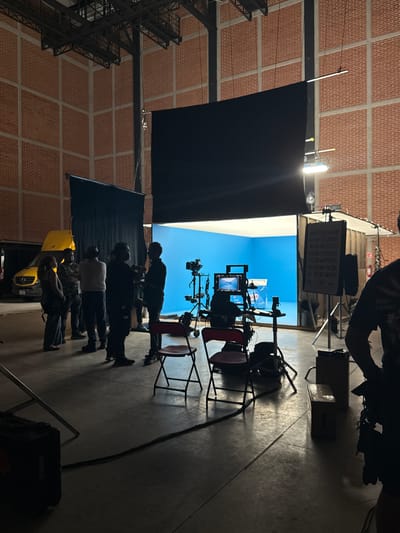 The image shows a film or television set in what appears to be a studio or large warehouse-like space in Tlalnepantla, Mexico, given the camera data. The location features exposed brick walls and a metal framework structure on the ceiling. A large black screen hangs from the ceiling, obscuring part of the set. A blue screen backdrop is visible in the background, illuminated by studio lights. In the foreground, several people are standing and observing the scene, silhouetted against the bright background. A yellow vehicle is partially visible near the group of people. Various pieces of filming equipment, including cameras and monitors, are positioned around the set, supported by tripods and stands. Several chairs are also present near the equipment.