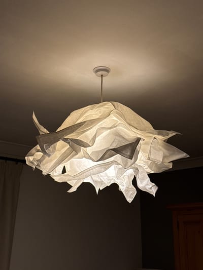 A low-angle, indoor shot shows a white, paper-like, abstract light fixture extending from a white wall. The fixture is mounted to the wall with a circular, white base and a silver rod. The light fixture itself is voluminous and irregular, with folded and layered material creating a sense of depth and texture. The light source within the fixture casts a warm, diffused glow, highlighting the folds and edges of the material. The wall behind the fixture is white, and there is a gradient of light cast on it. In the background, a dark area is visible, including the bottom corner of what appears to be a wooden cabinet door in the upper right corner of the image. There is no evidence in the image to indicate that the scene is located in Windermere, United Kingdom. No people or interactions are visible in the image.