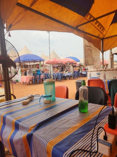 The scene depicts an outdoor market or event in Nibo, Nigeria, viewed from beneath a large, weathered orange and yellow canopy. In the immediate foreground, a table covered with a blue, white, and orange striped tablecloth holds a plastic water jug with a green lid, a small clear container with a blue lid containing a beige granular substance, a green plastic bottle with clear green liquid, a beige power strip, and a white power adapter with a black USB cable. A portion of a dark-colored standing fan is visible on the left. Beyond the foreground, numerous market stalls are established on reddish-brown dirt ground. These stalls utilize light-colored peaked tents and are shaded by large umbrellas, prominently blue and red. Multiple blue plastic tables and chairs are arranged in front of these stalls, indicating seating areas. A single individual wearing a blue shirt and a red cap is visible standing near a blue umbrella-shaded stall on the left. Additional indistinct figures are present further within the market. A tall utility pole rises centrally on the left. The sky overhead is overcast.
