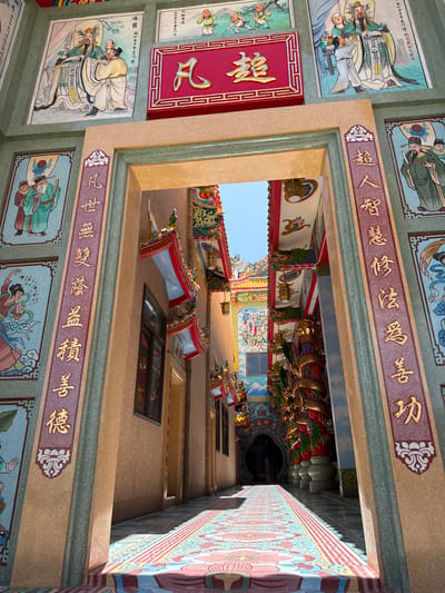 This image shows the exterior of a colorful, ornate temple in Ang Sila, Thailand. The foreground features a wide, arched entryway with intricate gold lettering on maroon panels on either side, framed by textured beige and green pillars. These pillars are adorned with traditional Chinese paintings and calligraphy. Through the entryway, a long, narrow walkway stretches into the distance, covered by a vibrant, patterned carpet with geometric and floral designs. The walkway is flanked by tall, red and gold pillars, decorated with dragon motifs. The architecture is elaborate, with brightly colored eaves and hanging lanterns visible on the temple's exterior. The sky is visible above the temple structures. The scene is well-lit, suggesting it was taken during the daytime, likely under clear skies.