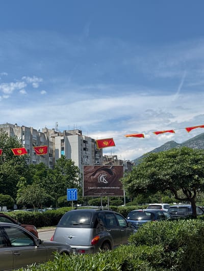 The image depicts a street scene in Bjeliši, Montenegro. Several Montenegrin flags are strung across the street. A building with a large advertisement for "Uskoro u ulici Vladimira Rolovica!" is visible. Cars are parked along the side of the street. The sky is blue with scattered clouds, and a mountain range is in the background. Trees and bushes line the street. A blue road sign is also visible.