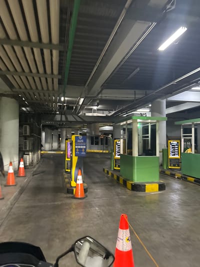 The image depicts an interior view of what appears to be a parking garage or a toll collection area, likely in Makassar, Indonesia. The scene is dominated by large, green automated toll collection booths, each equipped with digital displays showing currency symbols and numerical data. One display shows "Rp 0", indicating a zero balance or an initial state.

Several orange traffic cones are strategically placed on the concrete floor, some with reflective white and red stripes. One cone is positioned near a booth and is tethered by a thin yellow string, which also runs across the frame towards another cone further in the distance. A mirror, possibly from a motorcycle or scooter, is visible in the lower right portion of the image, suggesting this perspective is from a rider.

The overall lighting suggests it could be nighttime or an indoor environment with artificial illumination. The ceiling features long fluorescent light fixtures, and some sections of the walls are clad in dark, ribbed panels. The architectural elements, including elevated walkways and structural beams, are indicative of a multi-level parking structure or a similar facility.

The image seems to capture a moment of stillness, possibly during off-peak hours or before the toll collection system is fully operational. There are no visible people actively engaged in transactions, although a person in a yellow vest can be glimpsed inside one of the booths in a cropped view. The presence of the cones and the general layout suggests an operational area, possibly a payment or entry point. Text visible on a blue and yellow sign reads "SIAP KARTU FLUS-TUNAI" and "PRAKTIS".