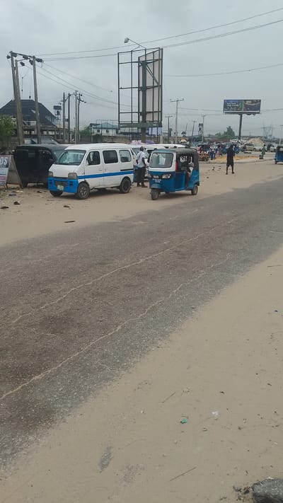 The image shows a street scene in Effurun, Nigeria. A white van with a blue stripe and a blue auto rickshaw (tuk-tuk) are prominent on the side of the road. Several people are standing near the van. The road is paved and shows signs of wear and tear. The shoulder of the road is sandy. In the background are other vehicles, people, utility poles, and a billboard structure. Overcast skies dominate the scene.