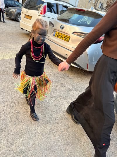 An image captured in Bnei Brak, Israel, depicts a young child dressed in costume, holding hands with an adult, walking on a paved surface. The child, with light-colored braided hair, has their face covered in black makeup with white streaks under the eyes, creating a distressed or crying appearance. They wear a black long-sleeved turtleneck shirt, a colorful hula-style skirt made of green, yellow, and red strips, patterned black tights, and dark flat shoes with buckles. Several strands of pink and yellow/gold beaded necklaces are draped around the child's neck. The child's left hand, also partially covered in dark makeup, is held by the right hand of an adult. The adult wears a brown long-sleeved top, a silver chain bracelet on their right wrist, and a long black skirt over dark shoes.
In the background, several vehicles are parked. A silver Toyota car (license plate: 220-81-802, yellow Israeli plate) is prominent behind the child. To its left, a white Mazda minivan (license plate: 40-731-69, yellow Israeli plate) is partially visible. The ground is a light gray, textured asphalt or gravel. Further in the background, outlines of buildings and parked cars are discernible, indicating an urban setting.