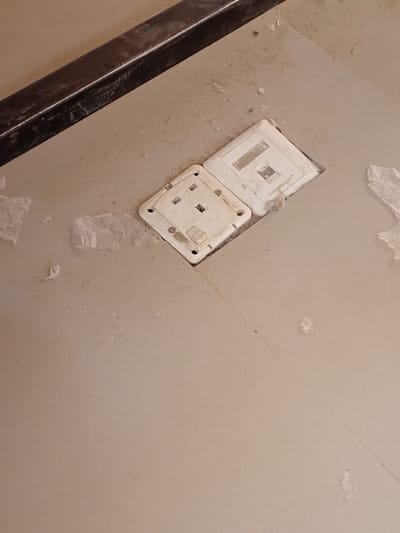 The image displays a close-up, downward-angled view of a dusty, light beige floor in an interior setting in Kwazailewa, Nigeria. Two white electrical fixtures are recessed into the floor. On the left is a square Type G electrical socket, secured by four screws, exhibiting visible dirt around its edges and in its pin receptacles. Adjacent to it on the right is a rectangular white cover plate, possibly for a data or communications outlet, also embedded in the floor and showing dust accumulation. The surrounding floor surface is covered in fine dust and scattered small, white, irregular debris fragments. In the upper left quadrant, a dark brown or black horizontal strip, appearing to be a baseboard or threshold, runs along the edge of the frame, also coated in dust. No people, active actions, or explicit interactions are visible.