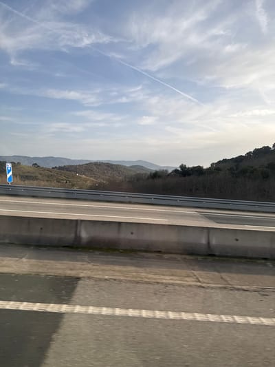 The image displays an elevated view of a highway or motorway in the region of Macedo de Cavaleiros, Portugal, under bright daylight. The foreground, shot from a moving vehicle, shows a dark asphalt road surface with a distinct dashed white lane marking. A low, grey concrete barrier, constructed from interlocking pre-cast sections, separates this foreground view from an elevated, multi-lane roadway. This elevated roadway features a lighter grey asphalt surface with multiple solid white lane markings. A silver metal guardrail runs along the left edge of this elevated road. Further left, partially visible beyond the guardrail, is a blue rectangular road sign with white pictograms, indicative of a service or amenity area (e.g., a fork and knife symbol). The background reveals a landscape of rolling hills and mountains, sparsely covered with brownish-green vegetation, suggesting a winter or early spring season. The sky is a light blue with scattered white clouds and a prominent, long white contrail from an aircraft extending diagonally across the upper left to the mid-right portion of the frame. Sunlight appears to be coming from the right, casting a shadow from the concrete barrier onto the lower foreground road. No individuals or other vehicles are distinctly visible in the immediate scene.
