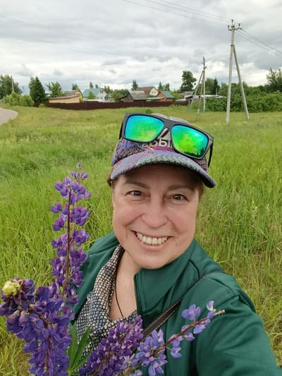 A woman is taking a selfie in a field of tall green grass under a cloudy sky. She is wearing a patterned baseball cap with sunglasses perched on top, a green jacket, and a patterned shirt visible underneath. She is holding a bunch of purple lupine flowers in her left arm. In the background, there are houses with various roof colors, a dirt road on the left, and utility poles with wires. The scene appears to be in a rural or semi-rural setting, possibly in the village of Gribchikha, Russia, given the style of houses and the natural landscape.