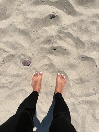 A top-down shot captures a pair of bare feet with white-painted toenails standing on a sandy beach. The feet are positioned in the lower center of the frame, with the ankles disappearing into the top of black leggings. The sand is light brown and appears to be made of fine grains, with subtle ripples and shadows indicating the texture and contours of the beach. A single, reddish-brown pebble with white speckles rests on the sand to the left of the feet. The scene is illuminated by bright sunlight, casting shadows on the sand. The location is identified as the city of Morskoye, Russia.
