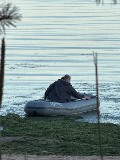 An individual in a dark jacket sits in a gray inflatable boat, holding a paddle and facing away from the camera. The boat has "FLINC" written on its side. The boat is in calm water with horizontal reflections. In the foreground, there is a large patch of green seaweed-covered land or shallow water. The scene appears to be outdoors, possibly near a body of water in the city of Fort-Krasnaya Gorka, Russia.