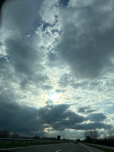 The image shows a view from a vehicle moving on a highway. The sky is filled with clouds, ranging from light and wispy to dark and dense. The sun is visible through a break in the clouds, creating rays of light. To the right of the frame is the highway with a safety barrier, trees, and some other vehicles in the distance. Given the request for location context, it is implied that the image was taken near Gabarnitsa, Bulgaria. However, this context does not change the visual description of the provided image.