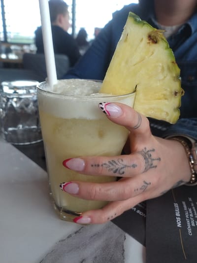 A tilted, medium-shot captures a hand holding a glass of yellow liquid, likely a cocktail, garnished with a pineapple wedge and a straw. The hand features tattoos on multiple fingers and decorative nail art. The drink is served over another glass. The background shows other individuals, furniture, and a menu with French text, partly visible with the words "NOS BULLES" and "CREMANT POLL FABAIRE." The location is identified as Schifflange, Luxembourg.