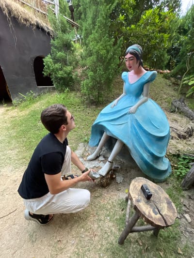 An outdoor scene, likely a themed garden or park in Nam Nha Trang, Vietnam, features a life-sized statue of a woman in a flowing light blue gown and a person interacting with it. The original image is rotated 90 degrees counter-clockwise.
The statue, possibly depicting a fairy tale character like Cinderella, has dark hair, a gentle expression, and bare feet. It appears to be leaning forward slightly, with its elaborate blue dress creating a sense of movement.
In front of and looking up at the statue is a person, likely male, wearing a black t-shirt, white pants, and black sandals. They are crouching or kneeling, holding a small, light-colored, possibly broken object in their hands, seemingly in the process of placing or presenting it to the statue's bare foot. Sunglasses are pushed up onto their head.
To the right of the statue, a small, rustic wooden table, made from a tree stump with three rough legs, holds a black action camera. The background is densely filled with lush green foliage and trees, creating a natural and verdant setting. In the lower left quadrant of the image, a dark opening, possibly an entrance to a cave or a structure, is visible. The scene is illuminated by natural daylight. No visible text is present.