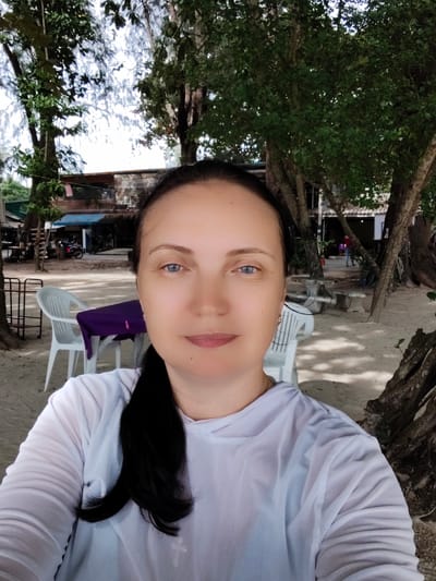 A woman is taking a selfie on a sandy beach in Sakhu, Thailand. She has dark hair pulled back into a ponytail and is wearing a white long-sleeved shirt. Behind her, there are trees, a building with a blue roof, and several white plastic chairs around a table covered with a purple cloth. The ground is sandy, and there is dappled sunlight filtering through the trees.