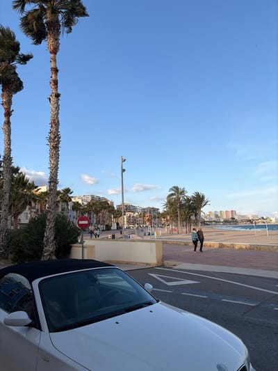 The image depicts a coastal street scene in Villajoyosa/la Vila Joiosa, Spain, under a clear blue sky with scattered white clouds. In the immediate foreground, the front hood and windshield of a white convertible car, likely a BMW Z4, with its black soft top deployed, are visible, angled slightly towards the right. White road markings, including an arrow and lane dividers, are visible on the asphalt in front of the car.
Beyond the vehicle, a beige-colored barrier separates the road from a wide promenade. Tall, slender palm trees with visible fibrous trunks line the left side of the scene and extend into the midground. Lush green foliage is present at the base of these trees. A red circular "No Entry" sign with a white horizontal bar is positioned near the left side of the promenade.
Further along the promenade, multiple streetlights are visible. In the midground, a row of multi-story buildings with varied, colorful facades (red, yellow, blue, white) lines the street. Two individuals are walking on the red-tiled promenade on the right side; one wears a teal-colored top and dark pants, the other a darker jacket and pants. Other figures are discernable walking further down the promenade in the distance.
To the right of the promenade lies a sandy beach area, where a beach volleyball net is visible. The Mediterranean Sea, with small waves, extends into the background. Distant buildings, including high-rises, stretch along the coastline under the bright sky, completing the urban coastal landscape.