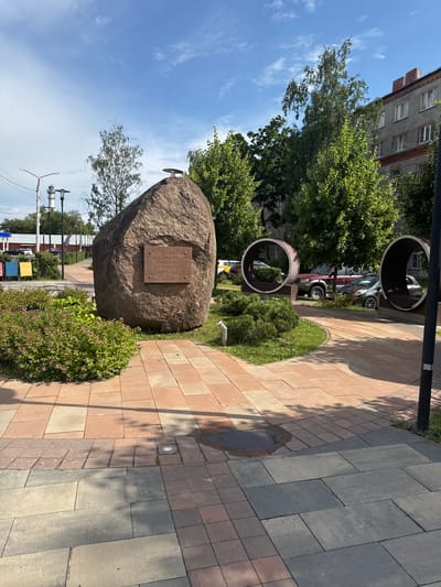 This image shows a public square in Ramenskoye, Russia, featuring a large boulder monument with a plaque, surrounded by a paved walkway and landscaping. The boulder is positioned centrally in the foreground, with a reddish-brown plaque attached to its face. The walkway is made of interlocking rectangular pavers in shades of red and gray. To the left of the boulder, there are lush green bushes with small pink flowers. In the background, there are several large trees, some with green foliage, and a multi-story apartment building. Two large metal pipe-like structures, possibly art installations or decorative elements, are visible to the right of the boulder. Several parked cars are visible behind the trees and installations. The sky is blue with scattered white clouds. The overall scene depicts a well-maintained urban public space.