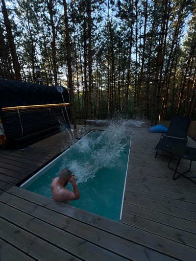 A man with a shaved head is partially submerged in a rectangular swimming pool, creating a large splash as he enters the water. He is facing away from the camera, with his arms raised to his face. The pool is surrounded by a wooden deck. To the left of the pool is a dark, barrel-shaped sauna with a chimney and an illuminated strip along its side. In the background, a dense forest of tall pine trees lines the horizon, with the sky visible through the branches. On the deck to the right of the pool, there is a black lounge chair and a small black table. A blue inflatable object is also visible on the deck. The scene appears to be in a natural, outdoor setting.