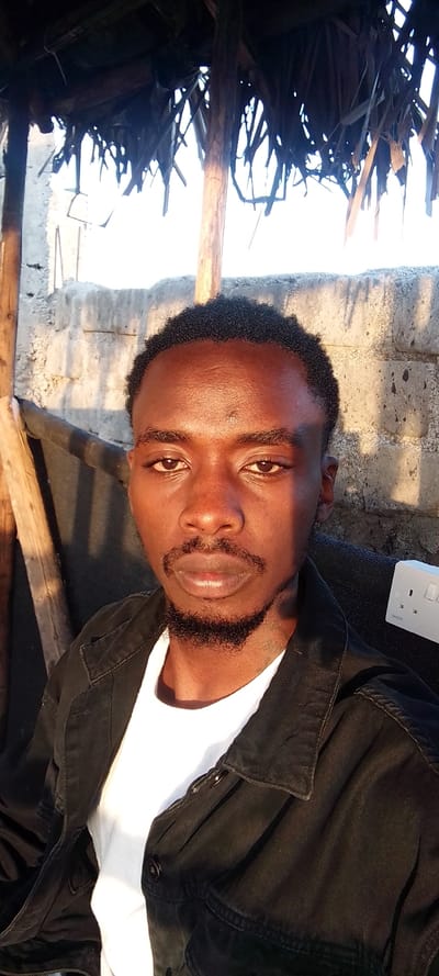 A male individual with dark skin, short dark curly hair, a mustache, and a beard is depicted in a front-facing portrait within Matathani ward, Kenya. He is wearing a white t-shirt beneath a dark, possibly black, button-up jacket. His gaze is directed forward. The background on the left features a rustic structure with rough wooden poles supporting a dark, dry thatch roof, and a visible concrete block wall. To the right of the subject, a white electrical wall outlet with two sockets is present against a dark background, possibly fabric or another surface. Strong natural light illuminates the scene, casting shadows, and suggesting an outdoor or semi-outdoor setting.