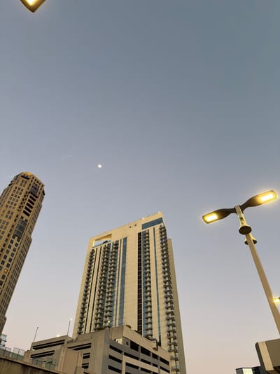 The image is a low-angle shot of the Atlanta skyline at dusk or dawn, featuring three buildings, a streetlight, and the sky. The building at the top right is a modern high-rise, primarily white with blue-tinted windows and numerous balconies. A second building at the bottom is older, constructed of yellow brick with a more ornate design. A third building on the right side of the frame is partially visible and appears to be a modern structure.
A streetlight with two illuminated lamps is situated in the upper part of the frame. The sky is a pale blue-gray, indicating twilight. A small, pale dot is visible in the sky which could be the moon or a star. The image appears to be taken from street level.