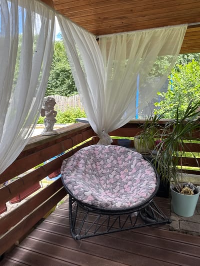 This is an eye-level, medium shot of a covered porch area on a sunny day, with the wooden floor of the porch filling the foreground. To the right, a black metal rocking chair with a round, cushioned seat covered in a pink and gray patterned fabric is centrally located. Behind the chair, to the right, are several potted plants on the wooden railing, including a tall, spiky plant in a light blue pot and another plant in a more decorative cream-colored pot. To the left of the chair, on the wooden railing, is a small, light-colored statue of a cherub or winged child. White sheer curtains are draped around the porch, obscuring some of the background view of lush green trees and a wooden fence. A fly is visible on the curtain in the upper left portion of the image. The overall setting appears to be a relaxed outdoor space.