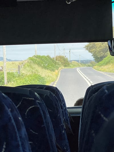 The image shows a view from the inside of a bus looking out at a winding road in Castlebane, Ireland. The foreground is filled with the backs of blue bus seats, some with white patterned stitching. In the middle ground, a paved road curves to the right, bordered by yellow and white dashed lines and surrounded by green grass and bushes. In the background, utility poles and wires are visible against a slightly overcast sky. Further in the distance, rolling hills and trees can be seen. A metal gate is also visible on the left side of the road. A person's head and shoulders, with brown hair, are partially visible between the bus seats.