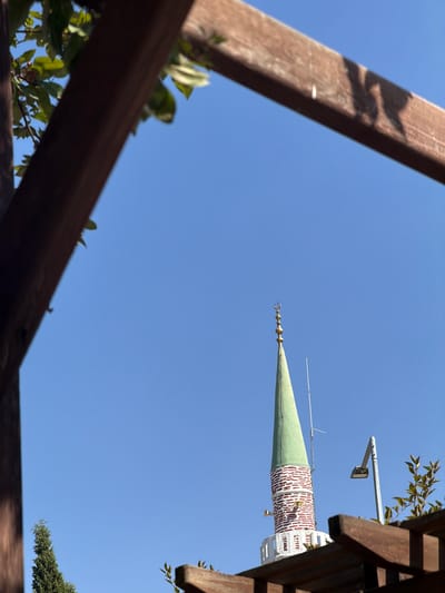 In Köprübaşı, Turkey, a minaret with a green conical spire topped by a crescent moon finial stands against a clear blue sky. The minaret's cylindrical base is adorned with a pattern of red brickwork interspersed with white sections. To the right of the minaret, a modern street lamp is visible. In the foreground, the edge of a brown wooden pergola partially frames the scene, with a few green leaves from an overhanging branch visible on the left. A tall, slender evergreen tree is situated in the lower left portion of the image, and the tops of more branches and wooden structures of the pergola are present in the foreground and right. The lighting suggests it is daytime.