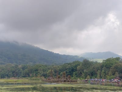 The image depicts a serene landscape at the Pura Ulun Danu Bratan temple complex, situated on the shores of Lake Bratan in Banjar, Indonesia. The sky is overcast with dramatic, grey clouds that partially obscure the lush green mountains in the background. The foreground is dominated by the placid waters of the lake, covered in a thin layer of green algae.

In the middle ground, the iconic Pura Ulun Danu Bratan temple complex stands majestically on a small island in the lake. Its distinctive tiered pagodas and intricate stone architecture are clearly visible. The temple appears to be constructed from dark stone and shows signs of weathering. Several small boats are moored near the temple.

Along the far shore of the lake, a bustling area with numerous colorful umbrellas and tents suggests a local market or gathering point. People are visible milling about, some walking and others gathered in groups, indicating tourist activity or local commerce. There are also several vehicles parked in this area.

The overall scene conveys a sense of tranquility and cultural significance, enhanced by the misty atmosphere and the natural beauty of the mountainous terrain surrounding the lake. The weather suggests a day that might be experiencing light rain or is about to.