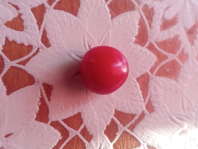 A close-up, top-down, medium shot shows a single, bright red, spherical object resting on a surface with a textured, light-colored, raised pattern. The pattern appears to be floral or lace-like, with interlocking shapes. The background surface is a reddish-brown, likely wood grain. The lighting creates highlights on the red object and casts subtle shadows. The overall scene is still and focused solely on the object and its immediate surroundings. No location in Semey, Kazakhstan is discernible.