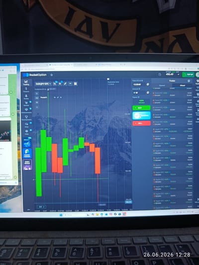 The image shows a laptop screen displaying the PocketOption trading platform. The platform shows a EUR/JPY OTC chart with candlestick patterns in green and red. There are also various trading interface elements such as expiration time, amount, payout, buy/sell buttons, and a list of recent trades. On the left side of the screen, a Telegram chat window is partially visible. The laptop keyboard is visible in the foreground with the date "26.06.2026 12:28" displayed on the screen. The background of the laptop screen displays a large, partially obscured logo with the letters "IAV". The overall scene appears to be an indoor setting, likely a home or office environment, with no specific identifiable Novovolynsk, Ukraine landmarks visible.