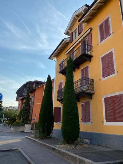 The image depicts a street scene in Milvignes, Switzerland, featuring a bright yellow building with red shutters and balconies. Adjacent to it is an orange building with a darker roofline. Two tall, slender cypress trees stand in front of the yellow building, positioned between it and the orange one. The sky above is blue with thin, wispy white clouds, possibly contrails. A paved walkway and a street are visible in the foreground, with concrete curbs. A blue circular traffic sign with a white symbol is partially visible on the left side of the image. The architecture suggests a European urban or suburban setting. There are no people or animals visible in the frame.