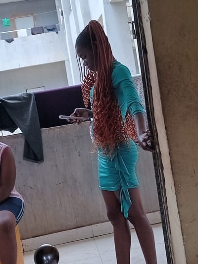 A young woman with orange braided hair, wearing a light blue dress, stands in a room. She is holding a mobile phone in her right hand. Behind her is a purple chair with a grey cloth draped over it. The floor is tiled. In the background, there's a doorway leading to another room with white walls and a shelving unit. Another person can be seen in the background wearing shorts. The room appears to be in Umueshi, Nigeria.