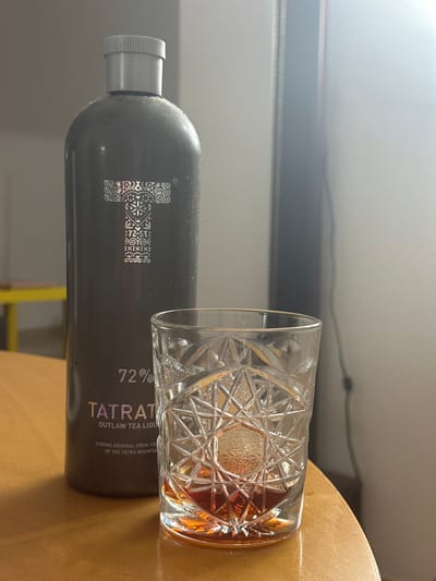 The media file presents an eye-level view of a bottle and a glass, placed on a light brown surface, likely a table, in Egkomi Lefkosias, Cyprus. The image appears to be rotated, with the items depicted as if standing upright.

On the left is a tall, dark-colored bottle with a metallic silver screw cap. The bottle's surface is matte and shows some faint dust particles. A prominent, stylized silver 'T' logo, composed of intricate patterns and symbols, adorns the bottle's side. Visible text on the bottle reads "72%", "TATRATEA", and "OUTLAW TEA LIQUEUR". Further text indicates it is "STRONG ORIGINAL FROM THE" "OF THE TATRA MOUNTAINS".

To the right of the bottle, a clear, heavy-bottomed glass is visible. The glass features an elaborate, cut-glass pattern with geometric shapes and starburst designs. It contains an amber-colored liquid, suggesting a spirit, liqueur, or a mixed drink. The background is a plain, light-colored wall or surface, with some subtle shadows and reflections indicating an indoor setting with ambient light. The composition creates a still life scene focused on the beverage items.